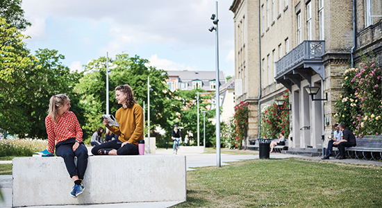 Two students sitting and talking in front of building at Frederiksberg Campus
